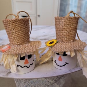 May include: Two scarecrow ornaments featuring white cylindrical bases with hand-drawn black eyes and mouths, orange triangular noses, and raffia hair. Each wears a burlap top hat, with one accented by a small orange plaid patch and one by a sunflower.