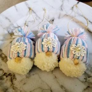 May include: Three small gnome ornaments with pink and blue striped hats, white pom-poms, and white lace flowers with pearl centers.