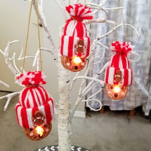 May include: Three Christmas ornaments hanging from a white tree. Each ornament features a red and white striped hat with a gingerbread man and a glowing light. The ornaments are suspended by gold string.