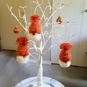 May include: A decorative white birch tree adorned with orange gnome ornaments and gold-colored baubles. The gnome ornaments have orange knit hats with pom-poms, white beards, and small wooden noses. The tree is lit with small white lights.