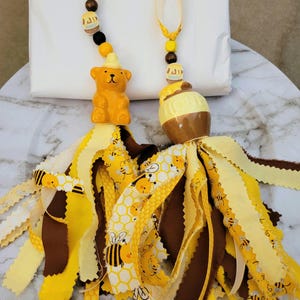 May include: Two decorative tassels with a honeybee theme. One features a ceramic bear, the other a honey pot. Both have yellow and brown fabric ribbons with bee and honeycomb patterns, and wooden beads.