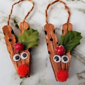 May include: Two handmade Christmas ornaments shaped like reindeer. Each ornament is made from a wooden clothespin with googly eyes, a red pom-pom nose, and a sprig of green leaves. A red and brown twine loop is attached for hanging.