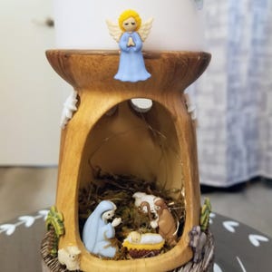 May include: A decorative nativity scene with a wooden-look base. The scene includes figurines of Mary, Joseph, baby Jesus, and an angel. The angel has yellow hair and a blue dress. The base has a hollowed-out center with a bed of hay and a string of lights.