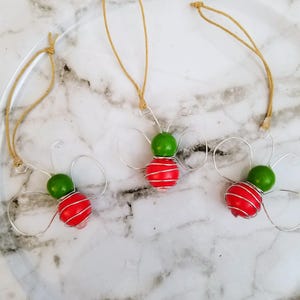 May include: Three handmade butterfly ornaments. Each ornament features a red, round bead wrapped with silver wire, topped with a green bead. The wings are crafted from silver wire, and they hang from a tan string.