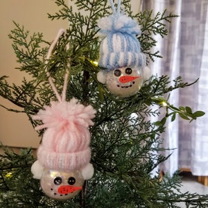 May include: Two snowman ornaments hanging on a Christmas tree. Each ornament is a clear ball filled with glitter, with a knitted hat and a pom-pom on top. One hat is light blue and white, the other is pink and white. The snowmen have button eyes and carrot noses.