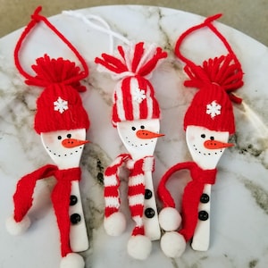 May include: Three handmade snowman ornaments crafted from wooden spoons. Each snowman features a red knit hat, scarf, and a carrot nose. The snowmen have black button eyes and a white pom-pom at the end of the scarf. A white string is attached for hanging.