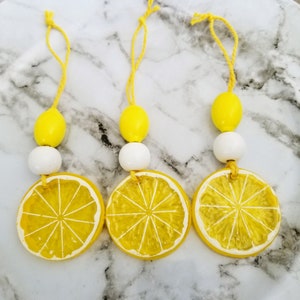 May include: Three yellow and white lemon-shaped ornaments with a string for hanging. Each ornament has a yellow lemon slice with a white center and a white bead on either side of the lemon slice.
