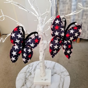 May include: Two decorative butterfly ornaments with patriotic star patterns in red, white, and navy blue. The butterflies are attached to a white decorative tree. Each butterfly has black antennae and a sparkling gem.