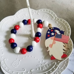 May include: A red, white, and blue beaded garland with a wooden gnome ornament. The gnome is wearing a red hat and holding a small American flag.