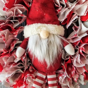 May include: A red and white Christmas wreath with a Santa gnome in the center. The gnome is wearing a red hat with white trim, a white beard, and red and white striped pants.