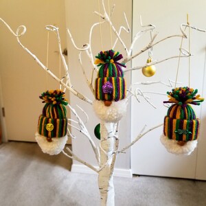 May include: Three handmade Mardi Gras gnome ornaments hanging from a white decorative tree. Each gnome has a knitted hat in purple, green, and gold stripes, a white beard, and a small decorative element. The ornaments are suspended by gold strings.