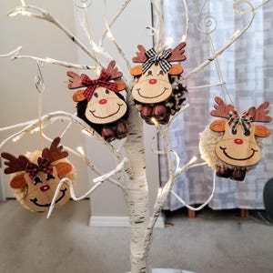 May include: Four reindeer ornaments hanging on a white birch tree with fairy lights. Each ornament features a wooden face, antlers, and a bow. The reindeer have different textures, including brown, black, and white fur.