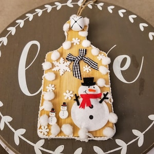 May include: A wooden Christmas ornament shaped like a cutting board, featuring a snowman, snowflakes, and a black and white checkered bow. The ornament has a jute string for hanging and is decorated with white pom-poms and small wooden snowflakes.