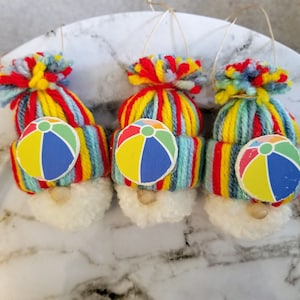 May include: Three yarn gnome ornaments with beach ball accents. The gnomes are wearing striped hats with red, yellow, blue, and green yarn. Each gnome has a white pom-pom beard and a beach ball with red, yellow, blue, and green colors.