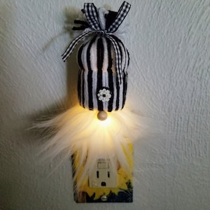 May include: A white and black striped gnome night light with a white fuzzy beard and a black and white checkered bow. The gnome is plugged into a standard electrical outlet.