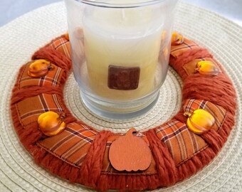 May include: A candle in a clear glass holder sits atop a decorative fall wreath. The wreath is made of orange yarn and plaid fabric patches, adorned with pumpkin-shaped embellishments. A wooden pumpkin cutout is also attached.