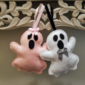 May include: Two felt ghost ornaments. One is pink with a pink ribbon and bow, the other is white with a black and white checkered bow. Both have black eyes and mouths. The ornaments are hanging.