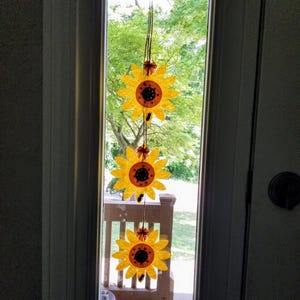 May include: A string of three felt sunflowers hanging in a window. The sunflowers are yellow with brown centers and are arranged vertically.