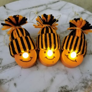 May include: Three orange, smiling face lights with black and orange striped knit caps and pom-poms. Each light has a small, bright bulb in the center of the face. The lights are connected by thin wires.