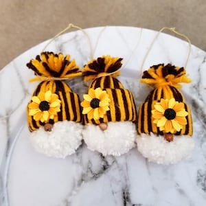 May include: Three handmade gnome ornaments. Each gnome has a white pom-pom beard, a brown and yellow striped hat, and a yellow sunflower. The hats have a yellow and brown pom-pom on top. Each ornament has a string for hanging.