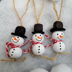 May include: Three wooden snowman ornaments with black top hats and red and white scarves. The snowmen have black eyes and a small orange nose.