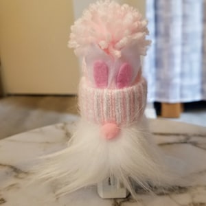 May include: A pink and white gnome with a fluffy white beard and a pink pom-pom hat. The hat has pink bunny ears and a white pom-pom on top.