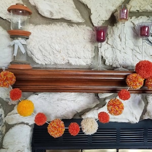 May include: A garland of orange, yellow, and white pom-poms hangs above a fireplace. The garland is made of yarn and is strung together with white thread. The fireplace is made of stone and has a black metal grate.