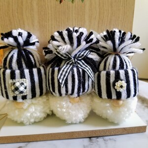 May include: Three black and white striped gnome ornaments with white pom-pom beards and black yarn hats. The hats have black and white checkered fabric accents and a black ribbon bow.