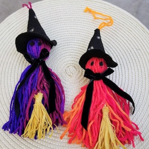 May include: Two handmade witch dolls with yarn hair in purple and red-orange hues. Each doll has a black witch hat with star accents, a black velvet bow, and a yellow straw broom. The dolls are decorative and festive.