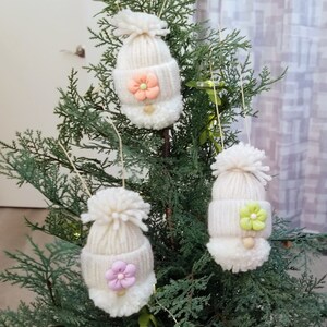 May include: Three white knitted gnome ornaments with pom-poms and felt flowers in pink, green, and light pink.