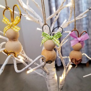 May include: Three wooden bunny ornaments with wire ears and ribbon bows. The bows are yellow and white, green and white, and pink. The ornaments are hanging from a white branch.
