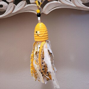 May include: A decorative yellow and white bee-themed tassel. The top is a yellow beehive shape with a painted bee. The tassel has yellow, white, and patterned fabric strips. A yellow ribbon with bee graphics and black beads complete the design.