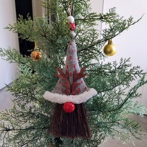 May include: A handmade Christmas ornament shaped like a gnome. The gnome has a gray knit hat with red accents, wooden antlers, a red nose, and a brown broom beard. The ornament is hanging on a green Christmas tree.