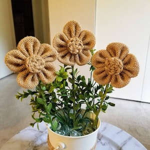 May include: Three burlap flowers in a white pot with green foliage. The flowers are brown and have a woven texture.