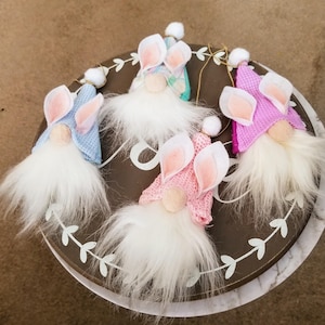 May include: Four Easter gnomes with white fur, wearing pink, blue, and light blue knit hats with bunny ears. The gnomes are sitting on a brown wooden surface with a leaf design.