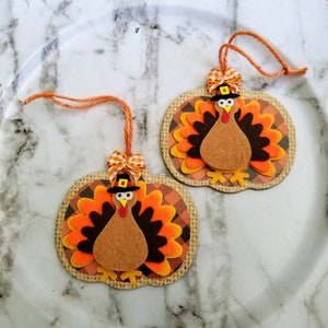 May include: Two pumpkin-shaped ornaments featuring felt turkeys with brown bodies, orange and black tail feathers, and black hats with yellow buckles. Each ornament has a burlap-textured border, a plaid background, and an orange twine hanger.