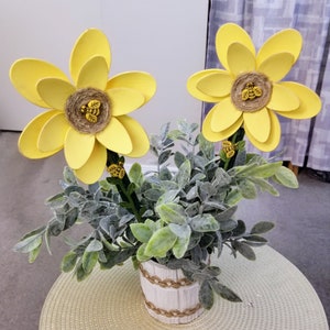 May include: Two yellow paper flowers with a bee design in the center are arranged in a white planter with green leafy foliage.