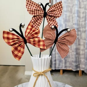May include: Three decorative butterfly stems in a white vase. The butterflies have red and yellow checkered wings, black pipe cleaner bodies, and pearl accents. The vase is tied with twine.