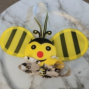 May include: A yellow and black bee-shaped ornament with a smiling face, red nose, and black eyes. The bee has large, transparent yellow wings with black stripes and a decorative bow. A small bee figurine is attached to the bow.