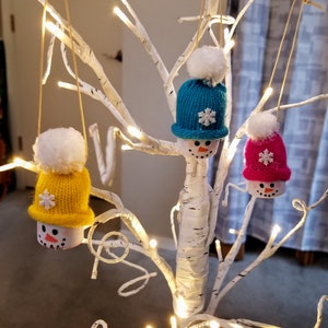 May include: Three snowman ornaments with knitted hats and white pom-poms hang from a white tree with lights. The hats are yellow, teal, and pink, each with a white snowflake. The snowmen have painted faces.