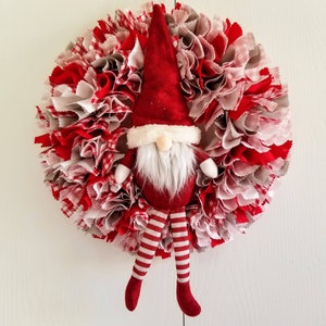 May include: A red and white fabric wreath with a red gnome with a white beard and striped legs hanging from it.