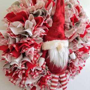 May include: A red and white fabric wreath with a gnome figure in the center. The gnome is wearing a red hat with a white trim, a white beard, and red and white striped pants.