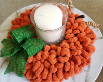 May include: A handmade orange yarn candle holder shaped like a pumpkin, with a green mesh bow and a white candle in a glass holder. The candle holder is on a white marble-patterned plate. A decorative item for fall.