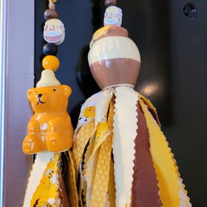 May include: Two decorative tassels with a honeybee theme. One features a ceramic bear, the other a honey pot. Both have fabric tassels in yellow, brown, and cream colors, with bee and honeycomb patterns.