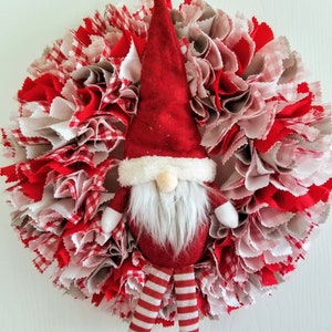May include: A red and white fabric wreath with a gnome figure in the center. The gnome is wearing a red hat with white trim, a white beard, and red and white striped pants.