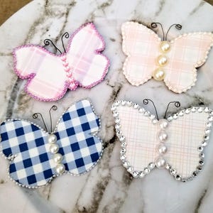 May include: Four decorative butterfly ornaments with different patterned fabric wings and pearl accents. The butterflies are pink and white, blue and white, and two variations of pink and white.