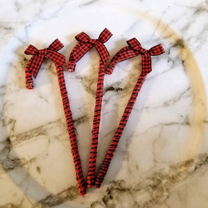 May include: Three decorative candy cane ornaments. Each cane is red and black checkered with a matching bow at the top. The ornaments are arranged on a white marble surface.