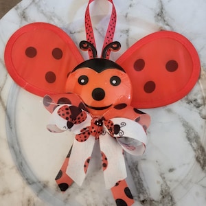 May include: A decorative ladybug ornament with red wings and black spots. The ladybug has a black head and a red ribbon for hanging. A bow with ladybug and polka dot patterns is attached to the front.