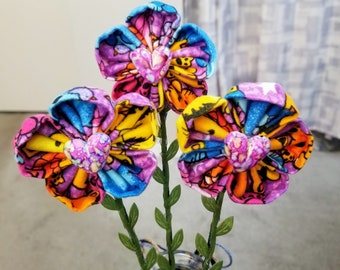 May include: A bouquet of three colorful fabric flowers with heart-shaped centers. The flowers are pink, blue, yellow, and purple. The flowers are arranged in a vase.