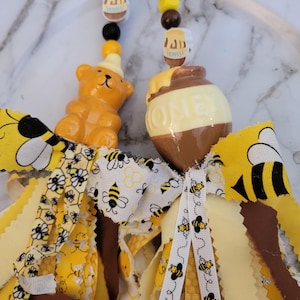 May include: Two decorative hanging ornaments featuring yellow and brown ribbons with bee and honeycomb patterns. One ornament has a honey bear bead, the other a honey pot bead with the word HONEY printed on it, accented by small wooden beads.